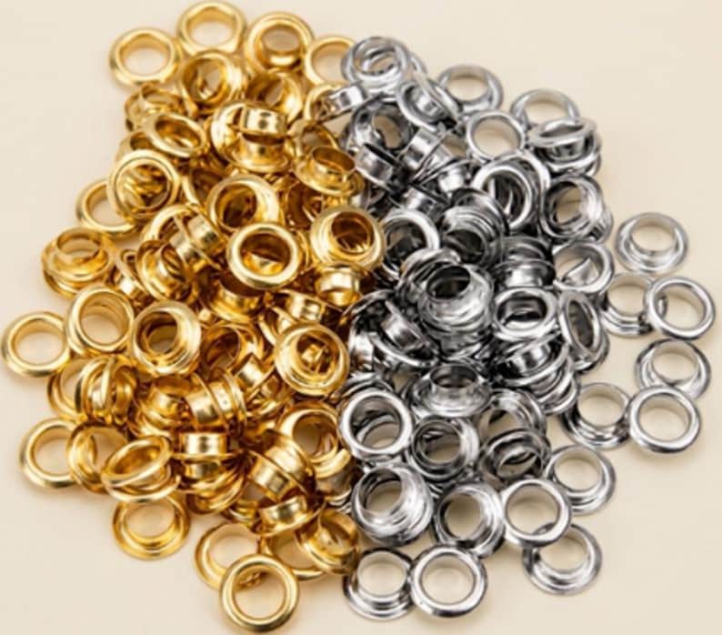 May include: A large collection of metal eyelets in two distinct finishes, featuring a shiny gold-tone color on the left side and a polished silver-tone color on the right side. These circular hardware components have a hollow center and a rim.