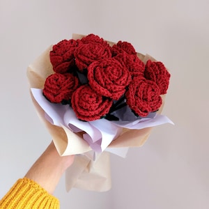 Puede incluir: Un ramo de rosas rojas de ganchillo envuelto en papel marr&oacute;n y blanco. Las flores hechas a mano tienen tallos de color verde oscuro. El ramo lo sostiene una persona que lleva un jersey amarillo.