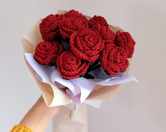 May include: A bouquet of red crocheted roses wrapped in brown and white paper. The handmade flowers are tightly wound, with dark green stems. The bouquet is held by a person wearing a yellow sweater.