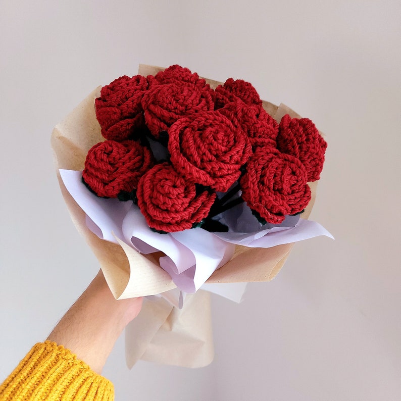 Puede incluir: Un ramo de rosas rojas de ganchillo envuelto en papel marr&oacute;n y blanco. Las flores hechas a mano tienen tallos de color verde oscuro. El ramo lo sostiene una persona que lleva un jersey amarillo.