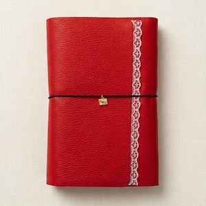 May include: A bright red journal with a textured surface, featuring a vertical strip of white lace trim along the right side. A thin black elastic band wraps horizontally around the cover, secured by a small gold-toned envelope charm.
