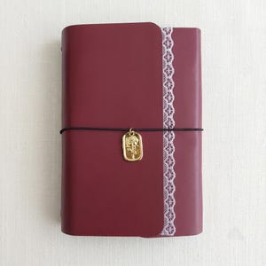 May include: A deep burgundy notebook with a smooth cover, featuring a vertical strip of white lace trim along the right edge. A thin black elastic band wraps around the center, holding a rectangular gold-toned charm embossed with a rose design.