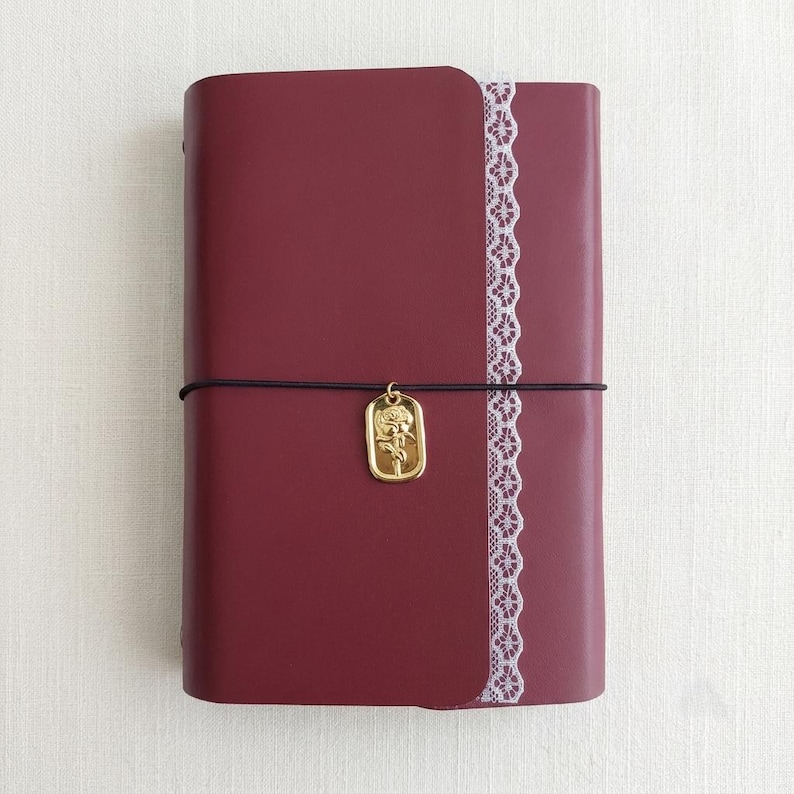 May include: A deep burgundy notebook with a smooth cover, featuring a vertical strip of white lace trim along the right edge. A thin black elastic band wraps around the center, holding a rectangular gold-toned charm embossed with a rose design.