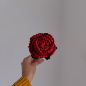 Puede incluir: Primer plano de una rosa roja tejida a mano con hojas verdes. La rosa est&aacute; bien enrollada, mostrando la textura del hilo. El art&iacute;culo artesanal se sostiene sobre un fondo liso, resaltando el color vibrante y la artesan&iacute;a detallada.