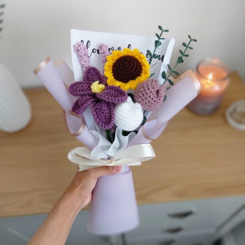 May include: A bouquet of handmade crochet flowers, including a sunflower, purple flower, and white flower, wrapped in lavender paper and tied with a cream ribbon. A card with the word "love" is visible. The bouquet is held in a lavender vase.
