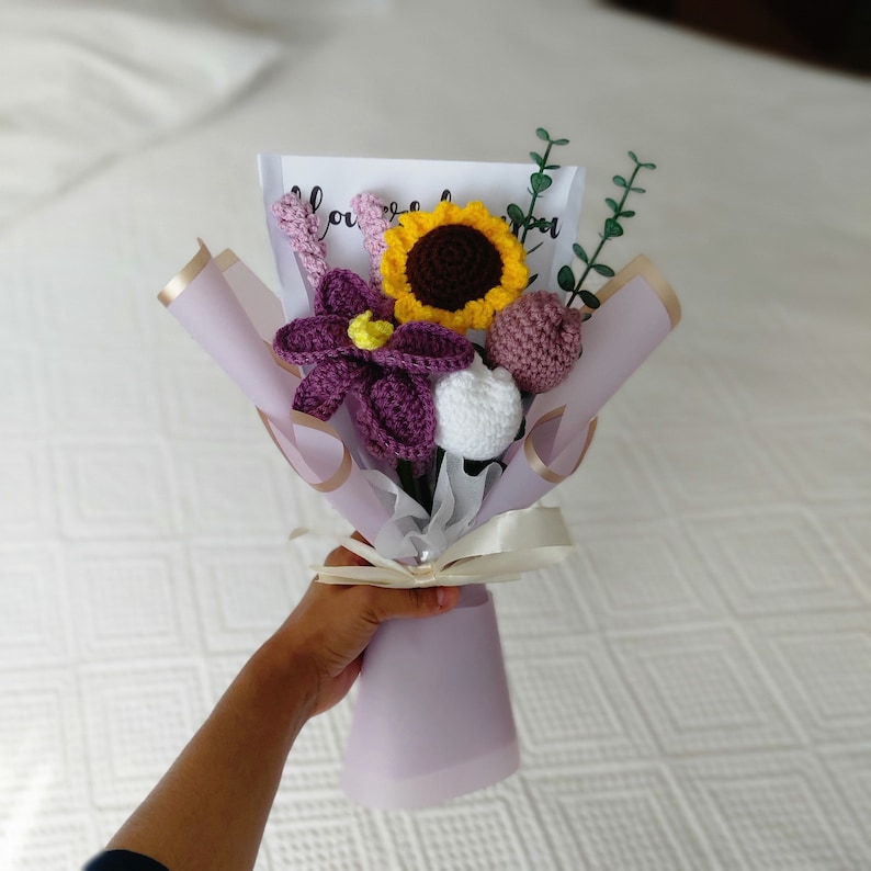 May include: A handmade crochet bouquet featuring a sunflower, purple flower, and other floral elements. The bouquet is wrapped in lavender paper and tied with a cream ribbon. The bouquet includes a card with the word "love" on it.