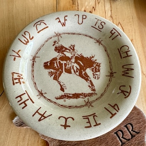 May include: A beige ceramic plate with a brown cowboy riding a horse graphic. The plate has a brown barbed wire border with the text "1KW2 F TP IS CH LAW JR 4T JE I A 44" around the edge.