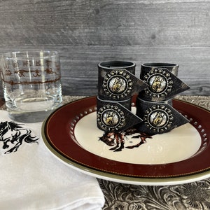 May include: Four black leather napkin rings with silver and gold horse head accents. The rings are stacked on a white plate with a red rim and a horse design. The plate is on a brown patterned surface.