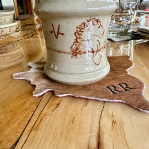 May include: A beige ceramic crock with a horse illustration and the letter "V" in brown. The crock sits on a cowhide-shaped coaster with "RR" branding. The table is made of wood.