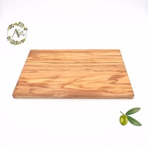 Peut inclure: Une planche à découper rectangulaire en bois d'olivier, de couleur brun clair et présentant un motif de grain de bois naturel. La planche a une surface lisse. L'image comprend également une branche d'olivier verte et un logo avec les lettres "A & C".