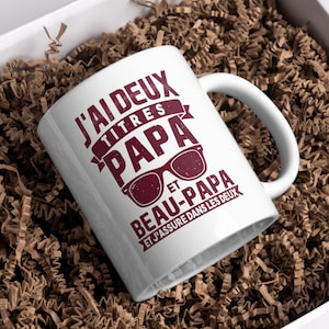 Può includere: Tazza in ceramica bianca con manico, con la scritta "J'ai deux titres PAPA et BEAU-PAPA" in bordeaux, insieme a una grafica di occhiali da sole. La tazza è presentata in una scatola bianca con imbottitura di carta marrone.