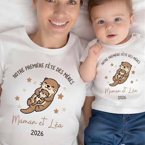 Peut inclure: Deux t-shirts blancs assortis avec un motif de loutre dessinée. Le motif représente une loutre embrassant un bébé loutre, entouré d'étoiles. Le texte "NOTRE PREMIERE FÊTE DES MERES", "Maman et Léa", et l'année "2026" sont imprimés.