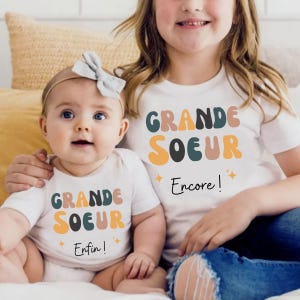 Peut inclure: Deux jeunes filles portant des t-shirts blancs avec les mots "GRANDE SOEUR" en lettres colorées. Le t-shirt de la plus jeune fille dit "Enfin!" et celui de l'aînée dit "Encore!". Les deux chemises ont des étoiles.