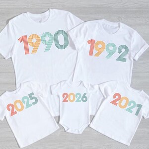 Peut inclure: Plusieurs t-shirts blancs avec des chiffres colorés rétro : 1990, 1992, 2025, 2026 et 2021. Les chiffres sont imprimés en rouge, orange, jaune, vert et bleu. Idéal pour une famille.