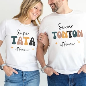 May include: Two white t-shirts with the text "Super TATA d'Amour" and "Super TONTON d'Amour" in a colorful, retro font. The shirts have a simple design with a star graphic. The people are wearing blue jeans.