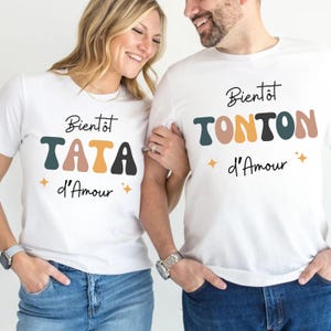 May include: White t-shirts with the text "Bientôt TATA d'Amour" and "Bientôt TONTON d'Amour" in black and colourful letters. The shirts have a retro font and star accents. The shirts are worn by two people.