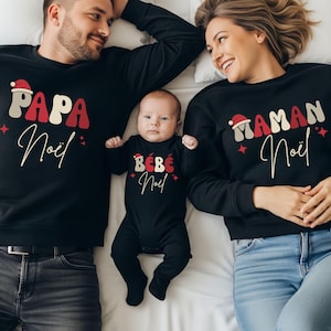 Op de afbeelding: Een familie in bijpassende zwarte kerstoutfits. De afbeelding toont een ouder en een kind die tops dragen met de woorden "PAPA Noël", "MAMAN Noël" en "BÉBÉ Noël" in rood, wit en beige. De outfits zijn feestelijk en op het thema van de feestdagen.
