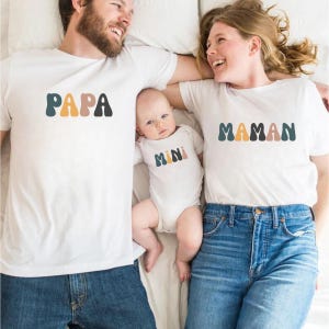Peut inclure: Une famille de trois personnes portant des t-shirts blancs assortis avec du texte coloré. Le t-shirt du père dit "PAPA", le t-shirt de la mère dit "MAMAN" et le t-shirt du bébé dit "MINI".