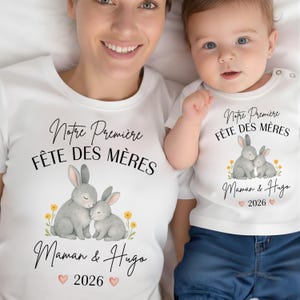 Peut inclure: Deux t-shirts blancs assortis. Chaque t-shirt affiche le texte "Notre Première Fête des Mères Maman & Hugo 2026" et une illustration à l'aquarelle de deux lapins gris entourés de fleurs jaunes. Ensemble assorti.
