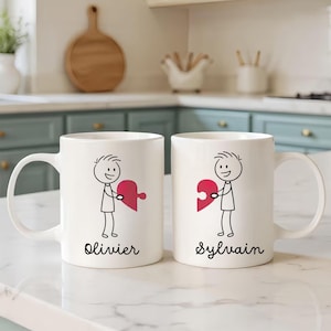 Op de afbeelding: Twee witte keramische mokken met een zwart stokfiguur dat een rood hartvormig puzzelstuk vasthoudt. Eén mok is gemarkeerd met "Olivier" en de andere met "Sylvain". De mokken staan op een wit aanrecht.