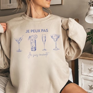 Peut inclure: Sweat-shirt beige avec des illustrations de cocktails bleus et le texte français "JE PEUX PAS Je suis enceinte". Le design présente un martini, un grand verre, une flûte à champagne et un verre à margarita.