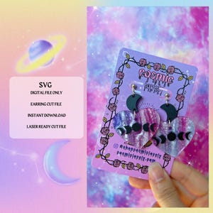 May include: A pair of heart-shaped earrings with a celestial theme. The earrings feature a marbled purple and pink design with black crescent moons and phases of the moon. The earrings are displayed on a purple card with the text "cosmic lovely".