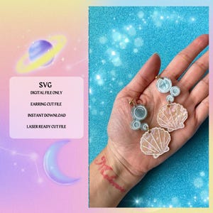 May include: A pair of earrings featuring a seashell and bubbles. The earrings are made of white and blue resin and have a gold-colored metal finding. The earrings are shown on a hand against a blue background.