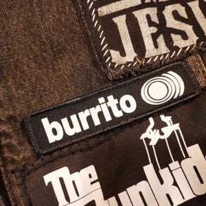 May include: Close-up of a dark denim jacket with several patches. One patch reads "burrito" in white text with a graphic of concentric circles. Other patches include the text "JESUS" and "The Junkids".