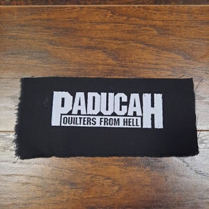 May include: A black rectangular patch with white text that reads "PADUCAH" above "QUILTERS FROM HELL". The patch has a frayed edge and is set against a wooden background.