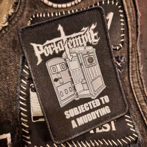 May include: Black rectangular patch with the words "Porto Temple" in white gothic lettering above a gray illustration of a portable toilet. The text "SUBJECTED TO A MUDDYING" is below the illustration. The patch is on a denim jacket.