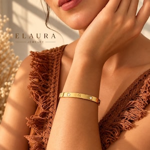May include: A gold-toned metal bangle bracelet featuring a smooth, polished surface with alternating circular screw-head motifs and small, clear, round-cut inset stones. The bracelet is worn on a wrist, with the text ELAURA JEWELRY visible nearby.
