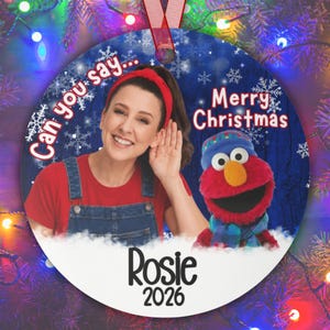 May include: A round Christmas ornament featuring a person in a red shirt and denim overalls with a red headband, alongside a red Elmo puppet wearing a blue hat and scarf. The text reads Can you say... Merry Christmas, Rosie, 2026.