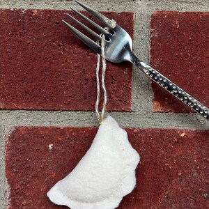 May include: A white felt pierogi ornament hanging from a silver fork against a red brick background. The pierogi has a scalloped edge and is suspended by a thin, off-white string. The fork's handle has a dotted pattern.