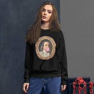 Puede incluir: Una sudadera negra con un retrato enmarcado en oro de un hombre soplando una burbuja rosa. El hombre lleva una chaqueta marrón y una camisa blanca.