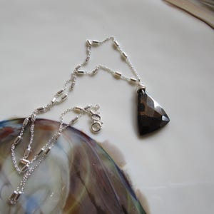 May include: A silver necklace with a geometric, dark brown pendant. The chain is delicate, with small, rectangular links. The pendant is a faceted, triangular shape. The necklace is displayed on a white surface with colorful, blurred accents.
