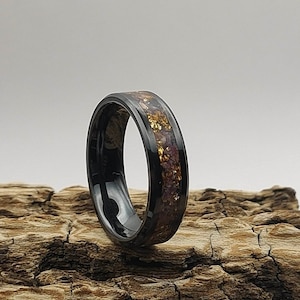 May include: A black tungsten carbide ring with a thin band of gold and purple inlay. The ring is resting on a piece of driftwood.