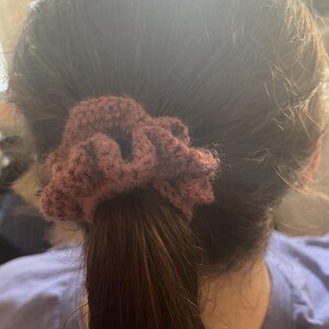 May include: A burgundy crocheted scrunchie secures a ponytail. The scrunchie is made of a textured yarn, and the hair is a dark brown color. The scrunchie is a hair accessory.