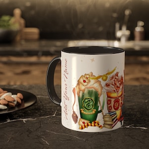 Può includere: Una tazza bianca e nera con un manico nero. La tazza ha uno sfondo bianco con un'illustrazione colorata di una bevanda magica con panna montata, un boccino d'oro e uno stemma di Hogwarts. Il testo "Add Your Name" è stampato sul lato della tazza.