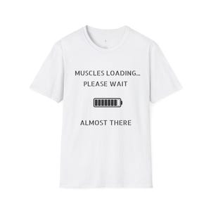 May include: A white short-sleeved t-shirt featuring black text that reads MUSCLES LOADING... PLEASE WAIT, followed by a battery icon graphic, and the text ALMOST THERE at the bottom. The design is centered on the front of the garment.