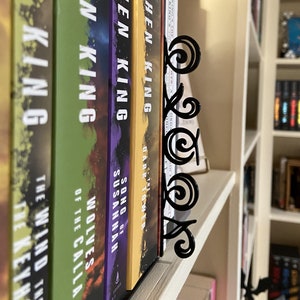 May include: A black metal bookend with a decorative scroll design holds a row of Stephen King books on a white bookshelf. The book spines are various colors, including green, purple, and yellow.