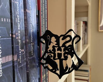 May include: A black metal bookmark in the shape of the Hogwarts crest, placed between the pages of a book. The bookmark features the Hogwarts school emblem with the letters "H" and "S" visible. The books are on a white bookshelf.