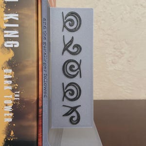 May include: A gray bookend with the word "BOOK" in a stylized font. The bookend is holding a book with a brown and orange cover. The bookend is made of a light gray material.