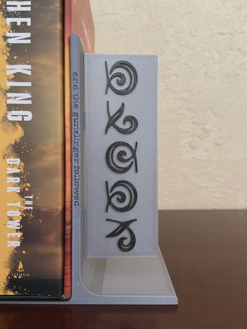 May include: A gray bookend with the word "BOOK" in a stylized font. The bookend is holding a book with a brown and orange cover. The bookend is made of a light gray material.