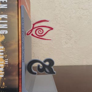 May include: A gray bookend with a black and red design. The bookend is holding a book with the title "The Dark Tower" by Stephen King. The bookend has a red eye design and a black ampersand symbol.