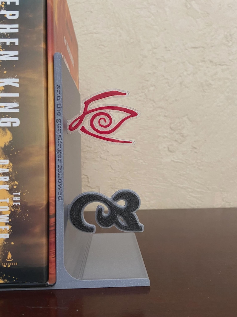 May include: A gray bookend with a black and red design. The bookend is holding a book with the title "The Dark Tower" by Stephen King. The bookend has a red eye design and a black ampersand symbol.