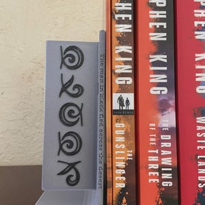 May include: A gray bookend with a stylized design and the word "OOK" in black. The bookend is holding up several books by Stephen King, including "The Gunslinger" and "The Drawing of the Three."