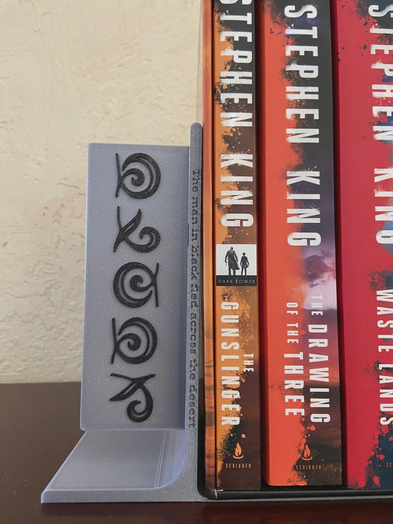 May include: A gray bookend with a stylized design and the word "OOK" in black. The bookend is holding up several books by Stephen King, including "The Gunslinger" and "The Drawing of the Three."