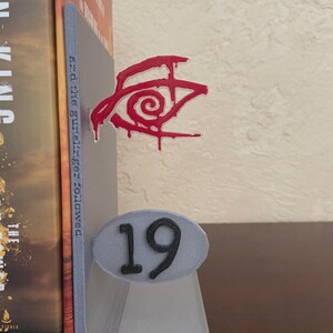 May include: A gray 3D-printed bookend with a red eye design and the number 19. The bookend is holding a book with a brown and gold cover. The eye design has a dripping effect.