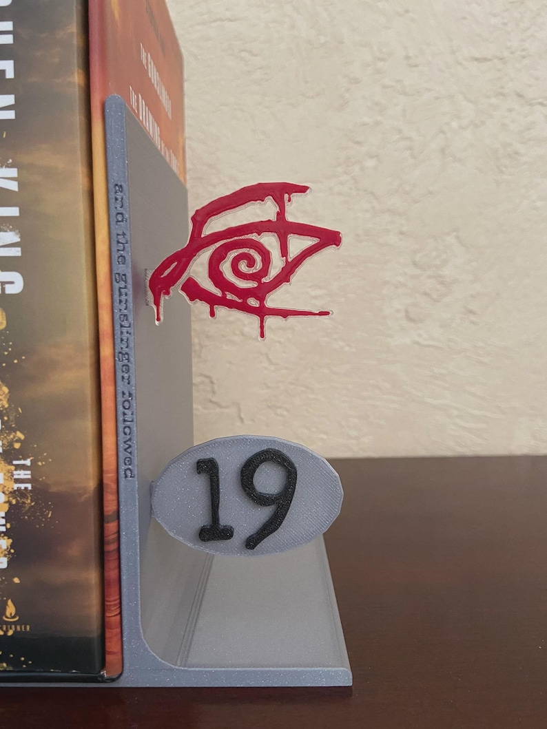 May include: A gray 3D-printed bookend with a red eye design and the number 19. The bookend is holding a book with a brown and gold cover. The eye design has a dripping effect.