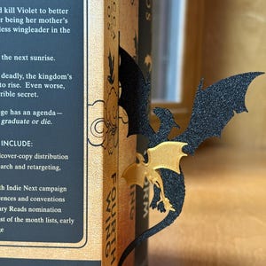 May include: A black and gold dragon bookmark is inserted into a book. The bookmark has a glittery texture and a detailed dragon design. The book's spine reads "Fourth Wing" and "Rebecca Yarros".